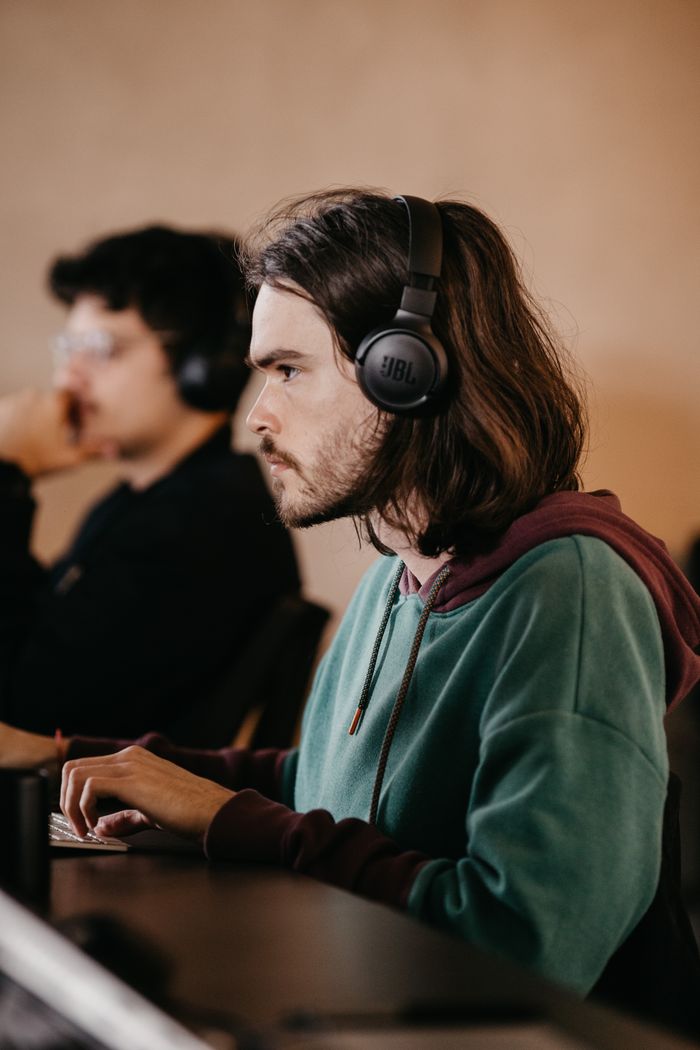 Young man with long hair working intently on a computer, wearing headphones, in an indoor setting.